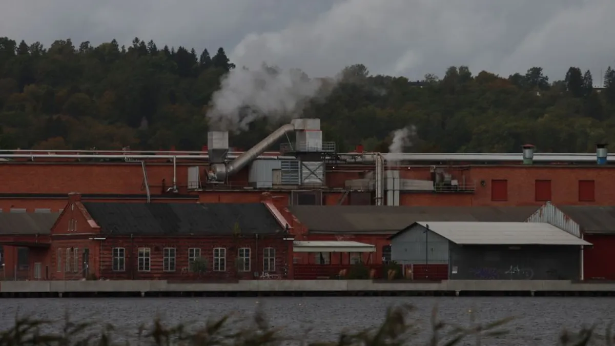 A factory with smoke emitting from chimneys along a river, surrounded by trees.
