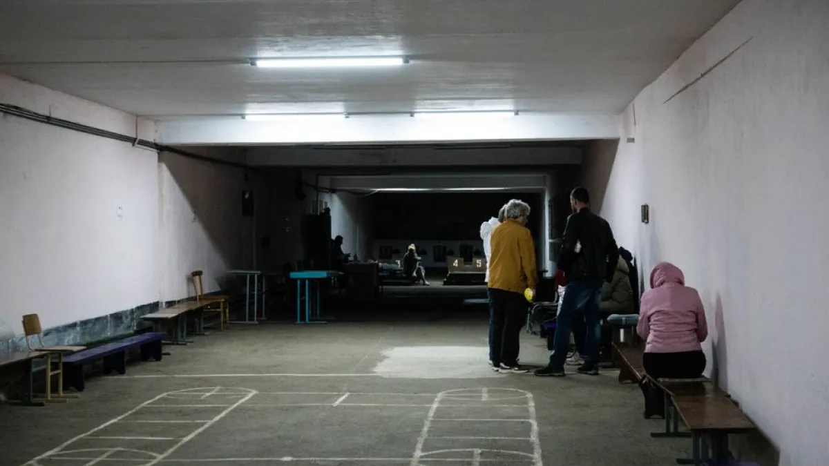 A group of people sitting and standing in an underground shelter with simple lighting and chalk drawings on the floor.