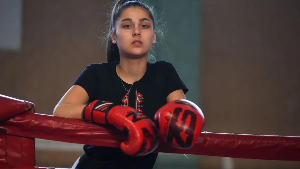 A young female boxer wearing red gloves rests on the boxing ring ropes indoors.
