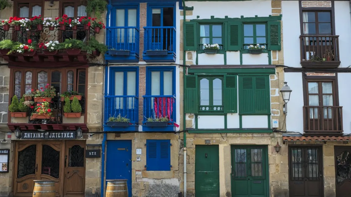 Colorful Traditional Spanish Houses in the Basque Country With Flowered Balconies in Coastal Village During Sunny Day - In a vibrant coastal village, rows of traditional Spanish houses showcase colorful facades adorned with flower-filled balconies. Each structure features unique architectural details, creating a picturesque atmosphere under a clear sky.