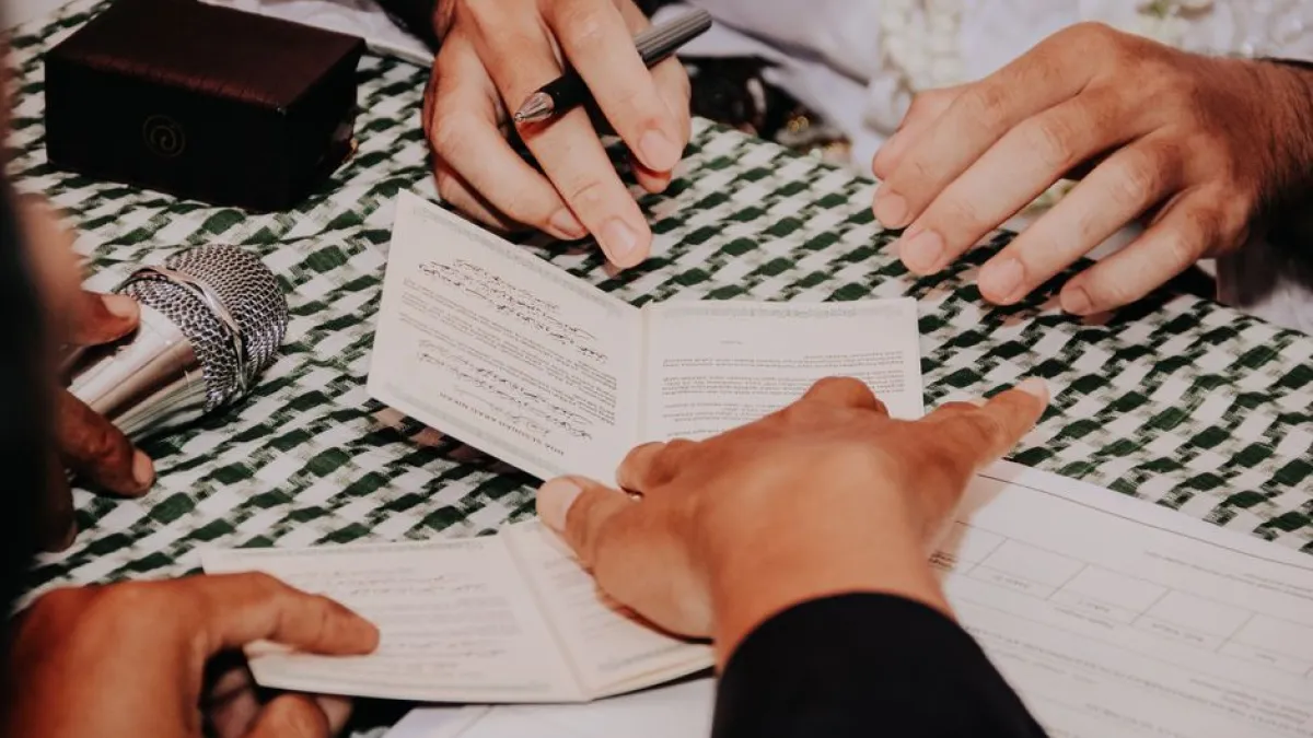 Multiple hands holding and signing a document during a formal ceremony with a microphone nearby.