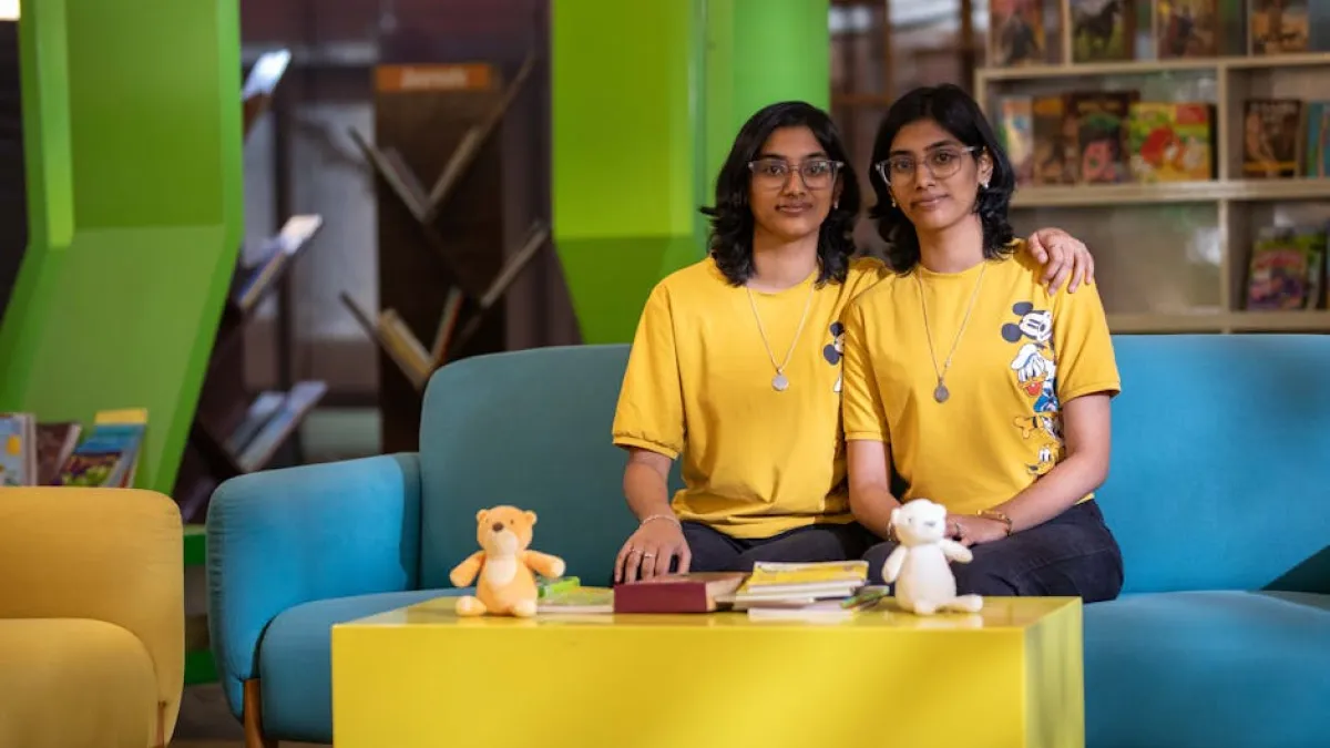 Twin sisters sitting on a colorful sofa in a library, smiling and wearing yellow t-shirts with cartoon prints.