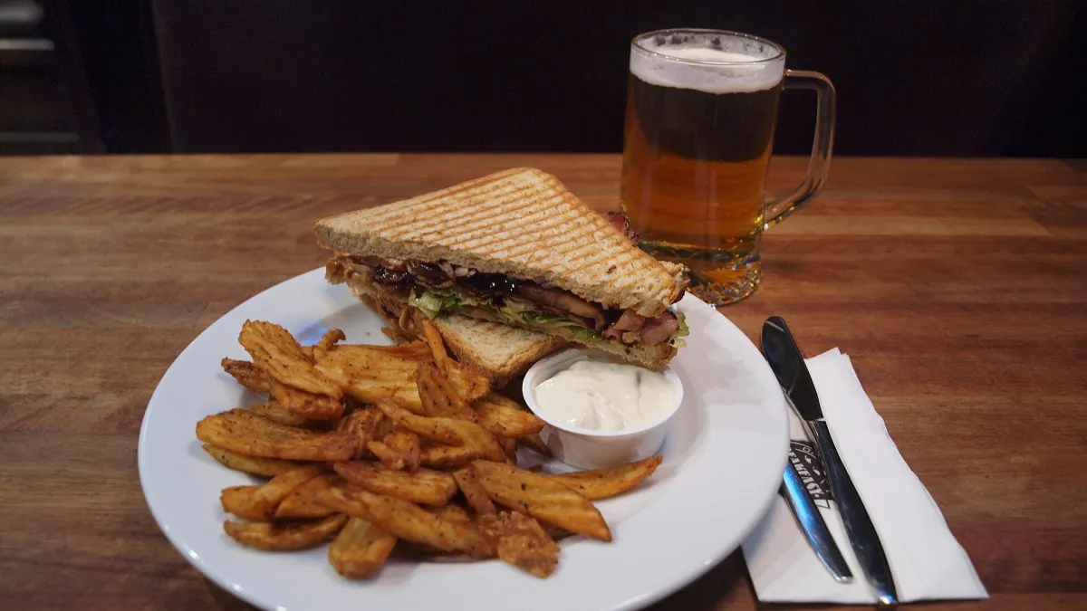 A smokey ham sandwich (called Ken "Smokey Ham" Eto alias "Tokyo Joe" on the menu), a toasted sandwich containing smoked bacon, salad, chipotle mayonnaise, tomatoes, onions and barbecue sauce, served with roast potatoes and dip sauce, along with a glass of beer (0.6 litres instead of the 0.4 to 0.5 litres usually available in Finland), served at restaurant Speakeasy in the Finlayson building complex in central Tampere, Pirkanmaa, Finland.